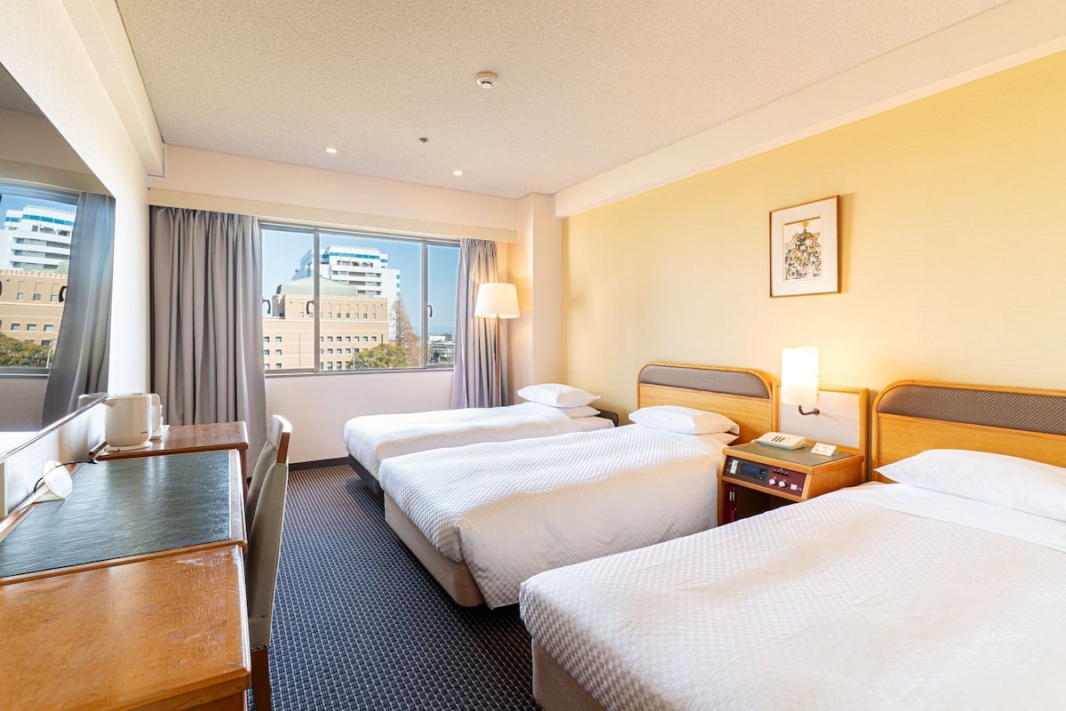 Une chambre d’hôtel avec deux lits, un bureau, une chaise, une lampe et une fenêtre donnant sur la ville.