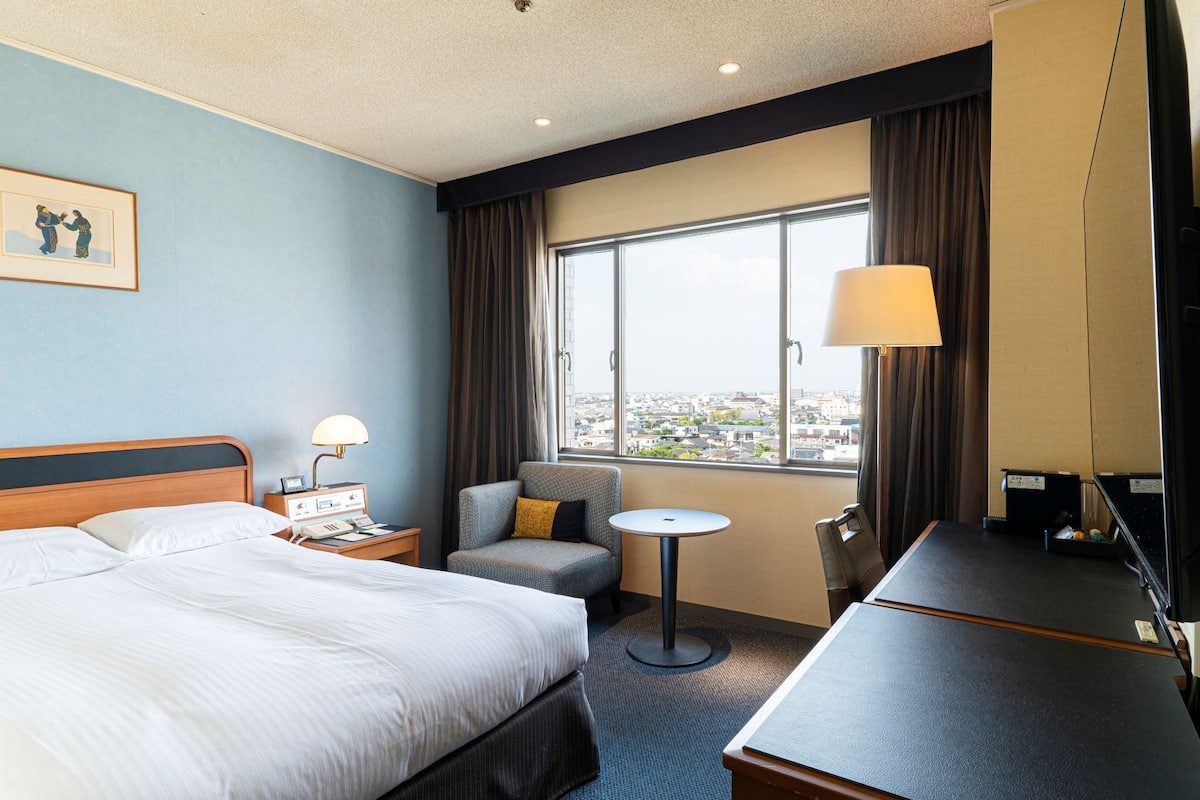Une chambre d’hôtel comprenant un lit, un bureau, une chaise, une petite table, une lampe, une fenêtre donnant sur la ville et un tableau encadré au mur.
