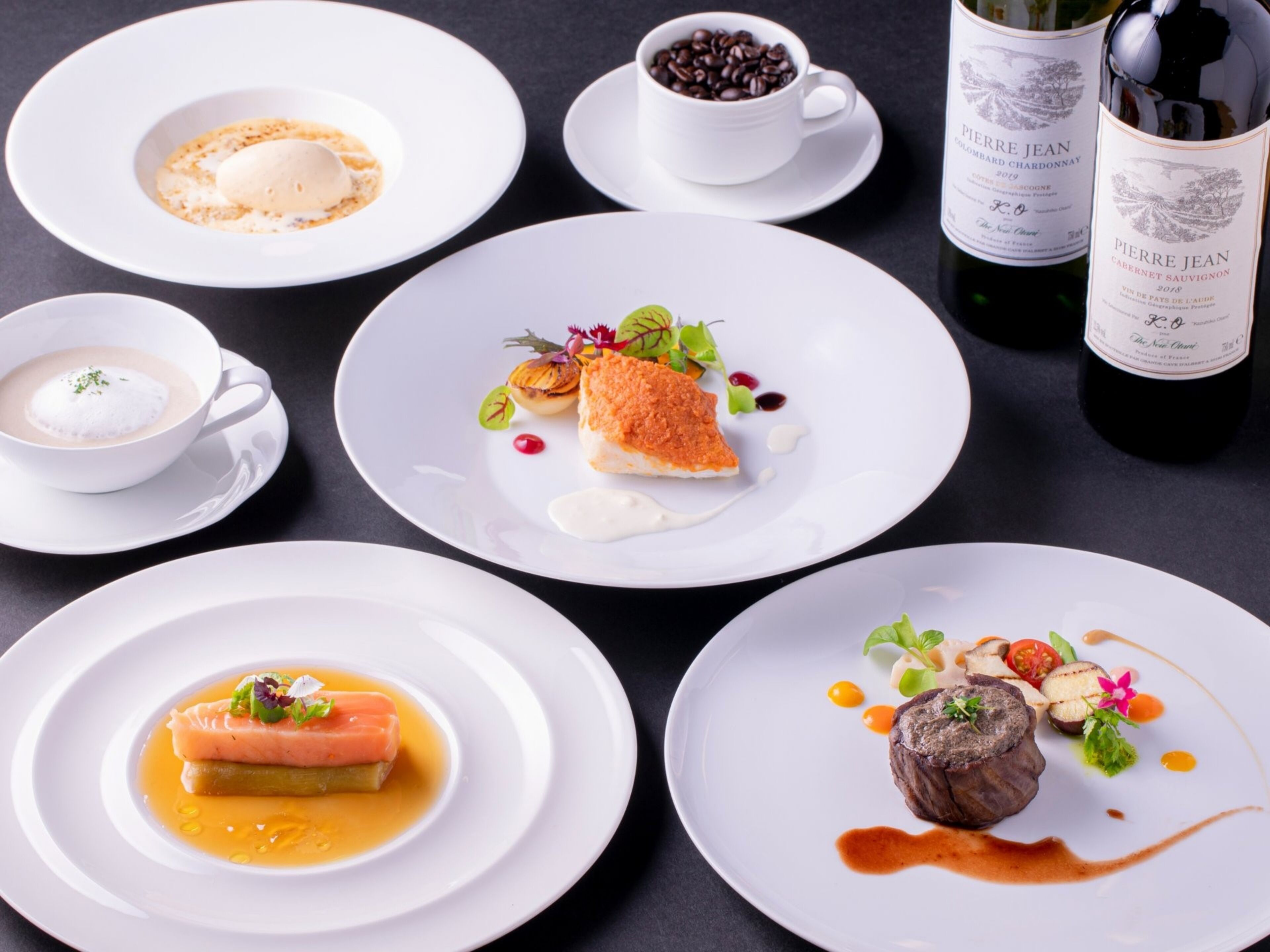 A variety of dishes on white plates, including seafood, a steak, and a dessert, with coffee and wine bottles in the background.