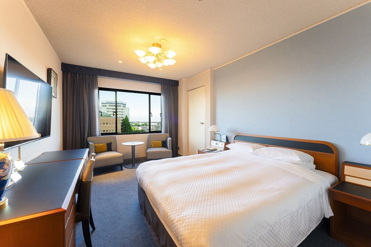 Une chambre d’hôtel avec un grand lit, un bureau, deux fauteuils, une petite table, une table de chevet avec une lampe et une fenêtre donnant sur la ville.