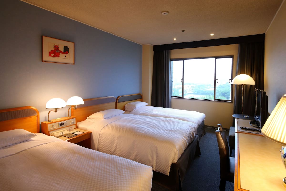 Une chambre d’hôtel avec deux lits, un bureau, une chaise, une lampe, un tableau au mur et une fenêtre donnant sur un paysage.
