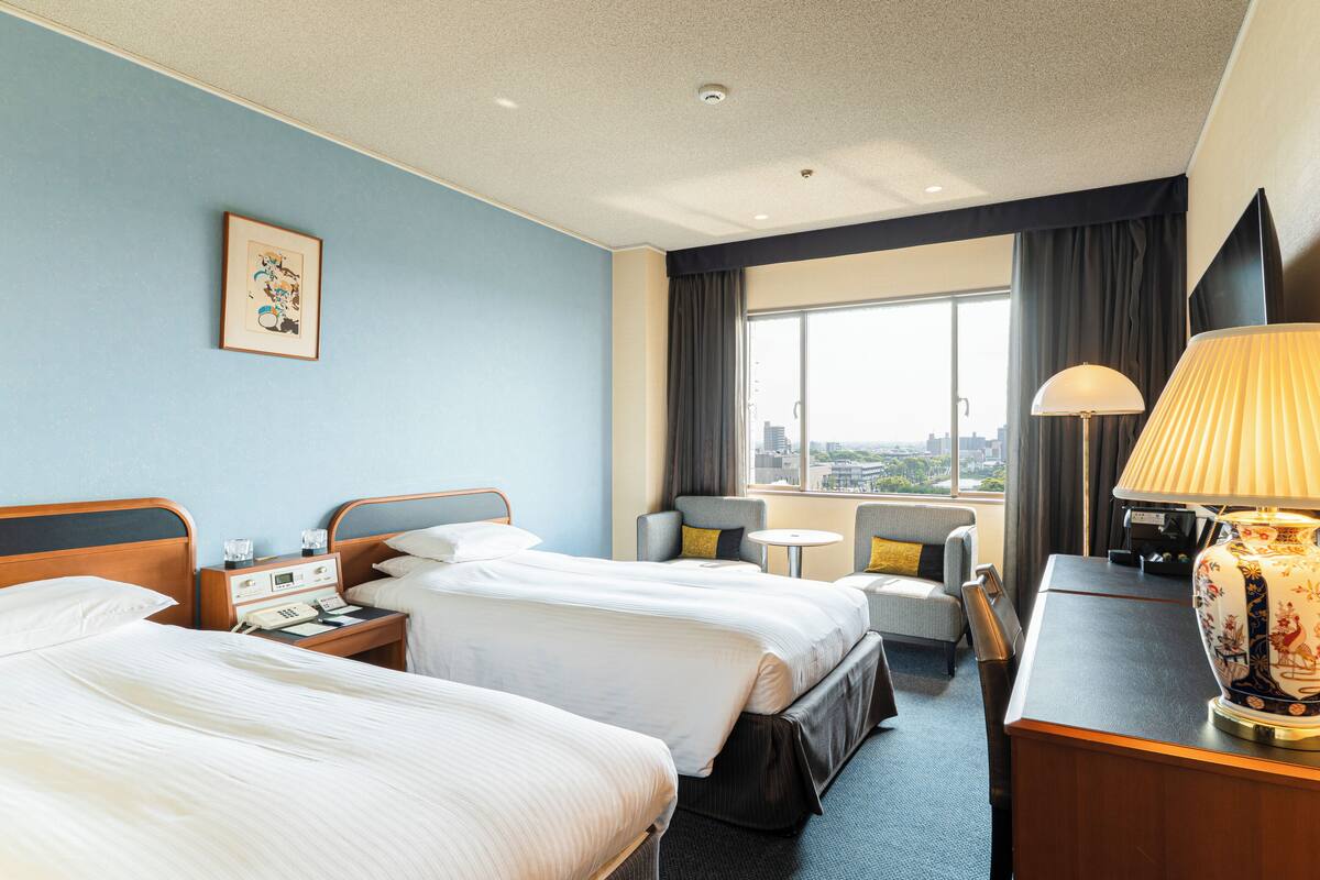 Une chambre d’hôtel avec deux lits, un bureau, une chaise, une lampe et une fenêtre donnant sur la ville.