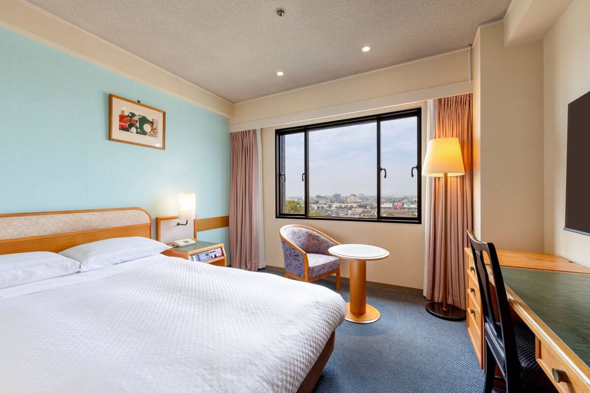 Une chambre d’hôtel équipée d’un lit, d’un bureau, d’une chaise, d’une petite table, d’une lampe, d’une télévision et d’une fenêtre donnant sur la ville.