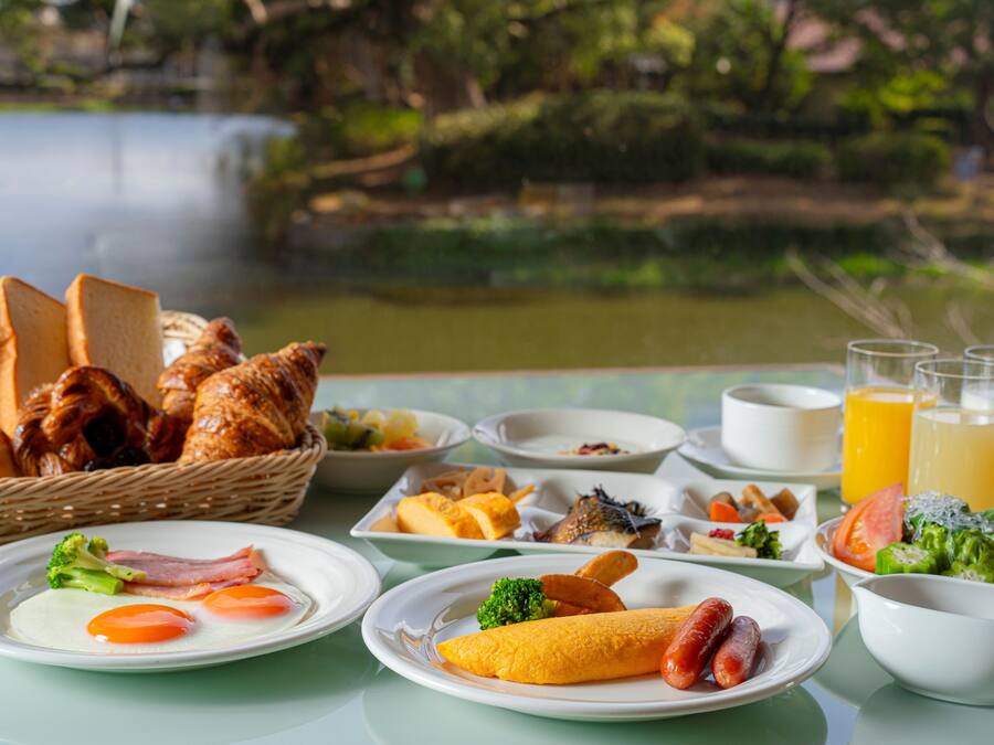 Un bufé de desayuno con diversos platos, como huevos, salchichas y pan, frente a una vista panorámica de un lago.