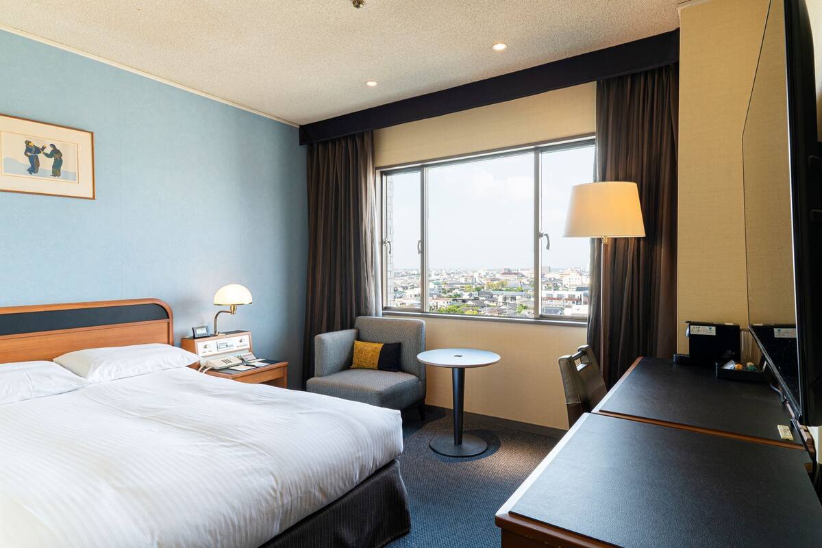 Une chambre d’hôtel équipée d’un lit, d’un bureau, d’une chaise, d’une petite table, d’une lampe et d’une grande fenêtre donnant sur la ville.