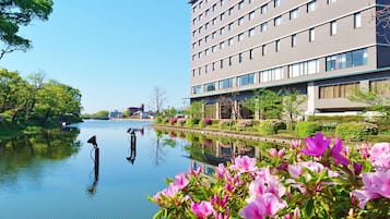 A modern building with a reflective water body in front, surrounded by lush greenery and flowering plants.