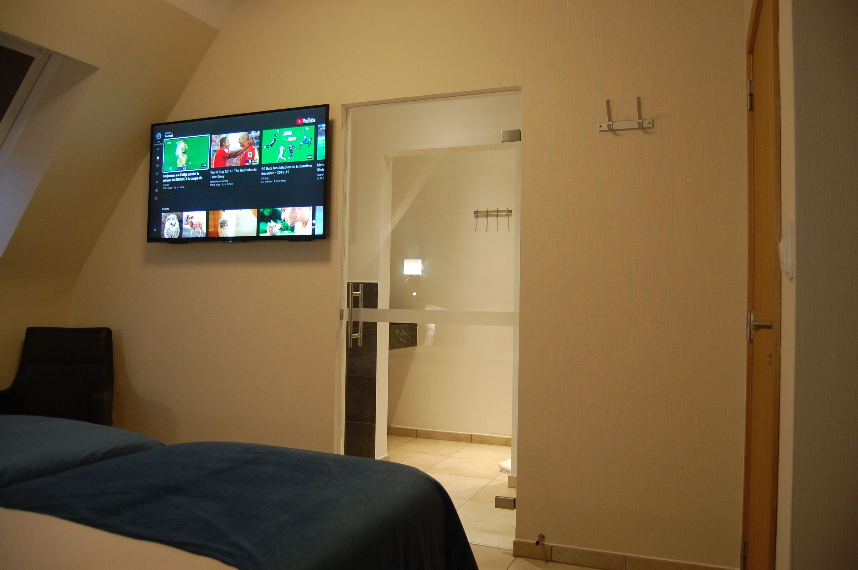 Un dormitorio con cama, televisor montado en la pared mostrando varios canales, y un baño visible a través de una puerta abierta.