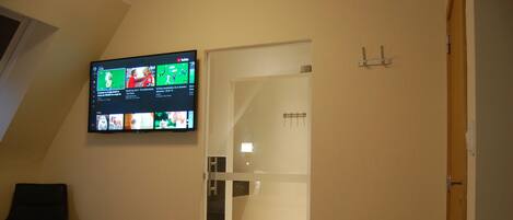 Un dormitorio con cama, televisor montado en la pared mostrando varios canales, y un baño visible a través de una puerta abierta.