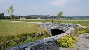 Un ponte di pietra sopra un prato erboso, con un sentiero che conduce a una costruzione in pietra.