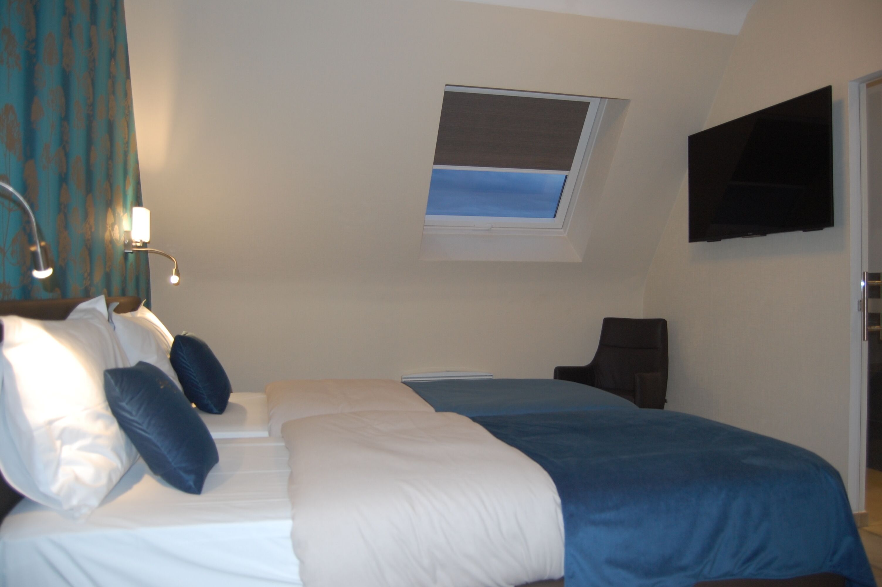Quarto de hotel com cama, poltrona, televisão, janela e cortina.