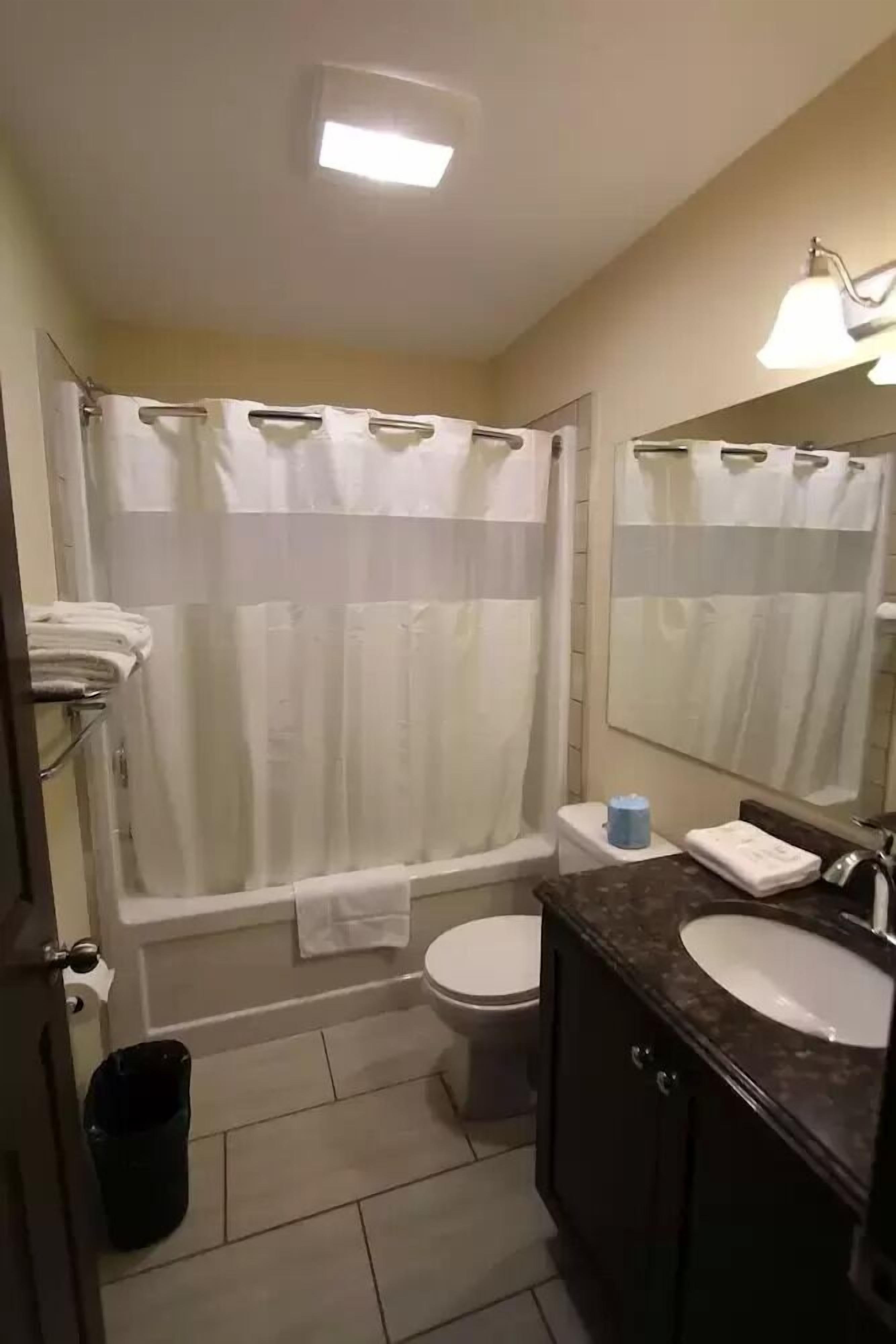 Un baño con bañera, inodoro y lavamanos. Hay un perchero con toallas, un cubo de basura y un espejo sobre el lavamanos.
