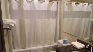 Un baño con bañera, inodoro y lavamanos. Hay un perchero con toallas, un cubo de basura y un espejo sobre el lavamanos.