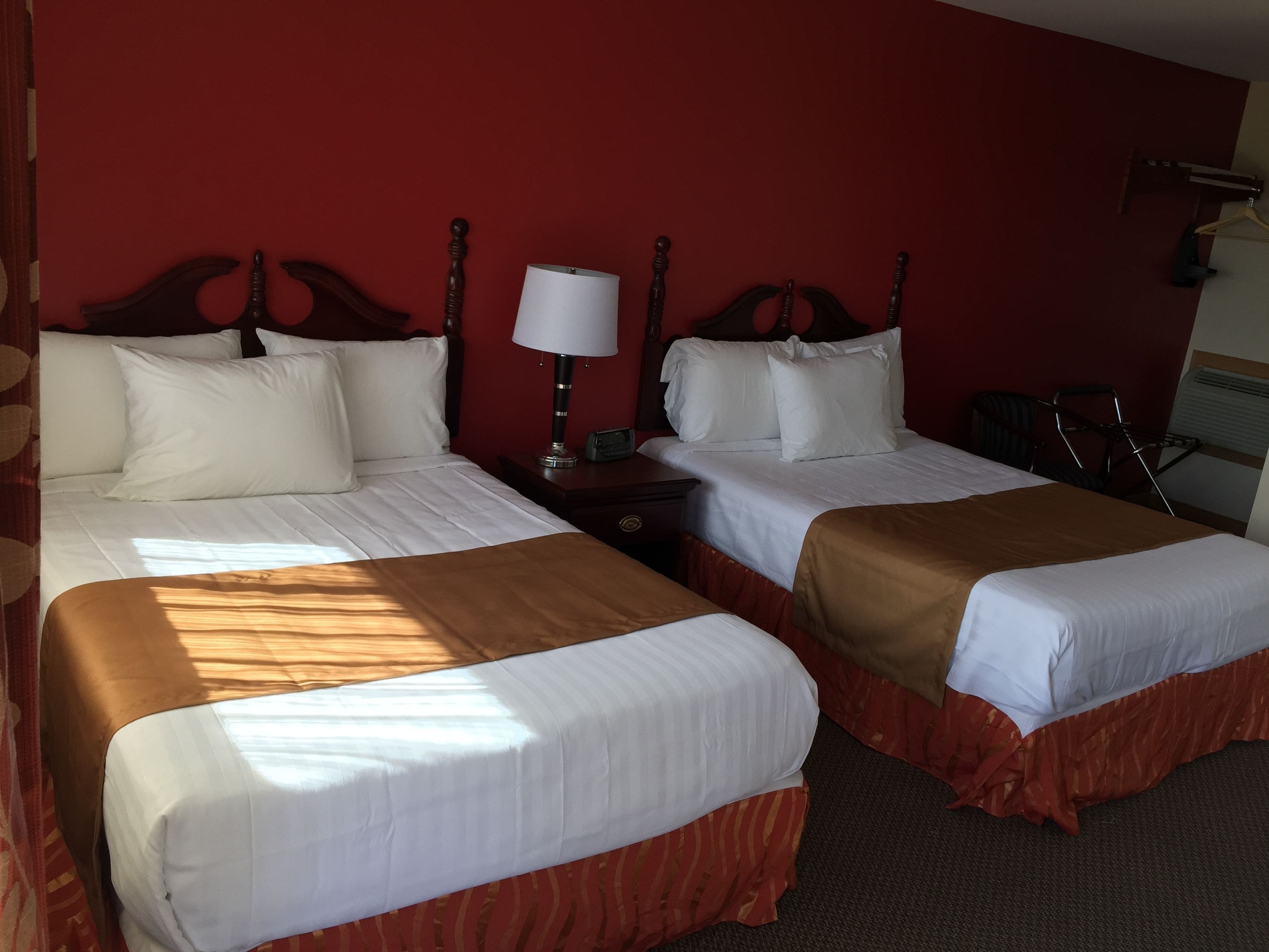 Habitación de hotel con dos camas, cada una con ropa de cama blanca y cabeceros marrones. Entre las camas, hay una mesita de noche con una lámpara y un teléfono.