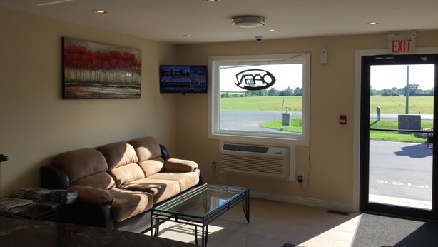 A lobby with a brown leather sofa, a glass-top coffee table, a painting, a television, a window with a view of a road and fields, and an exit door.
