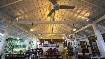 Een restaurant met een hoge plafond, houten balken en een centrale plafondventilator. Tafels en stoelen zijn er voor het diner gezet.