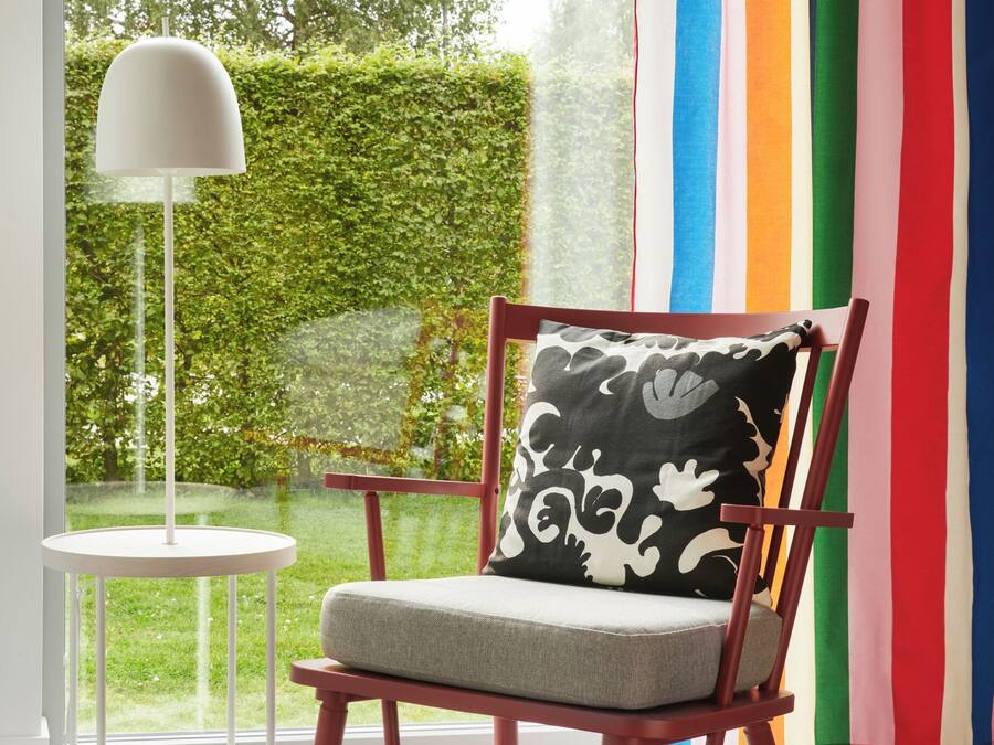 A chair with a patterned cushion, a white floor lamp, and a view of a green hedge through a window.