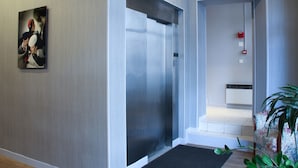 Um elevador moderno com acabamento metálico, um detector de incêndio fixado na parede e uma obra de arte emoldurada acima.