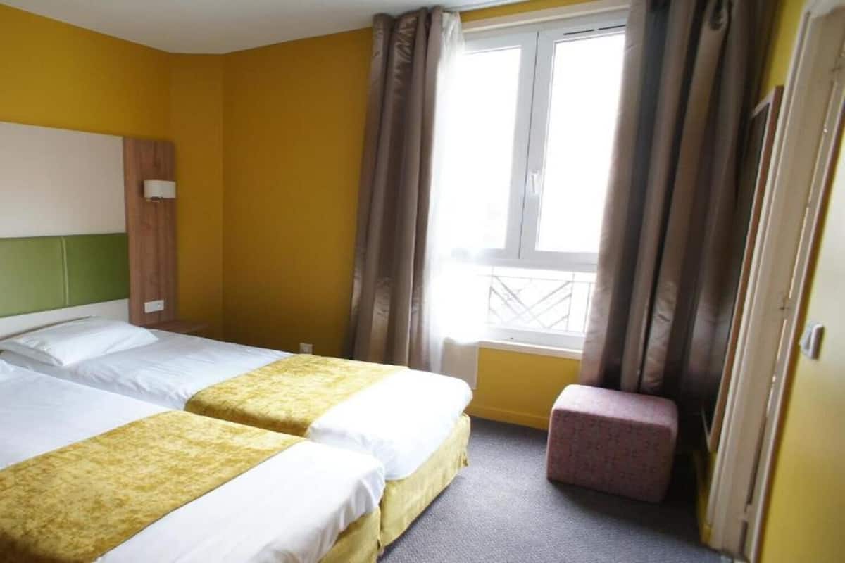 Habitació d'hotel amb dos llits, una finestra amb cortines, una petita banca i un capçal de llit.