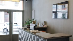 Balcão de recepção com divisória xadrez, uma planta em vaso, um quadro e uma janela com vista para a rua e carros estacionados.