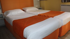 Uma cama de casal com roupa de cama branca e laranja, uma mesinha de cabeceira e uma luminária fixada na parede.