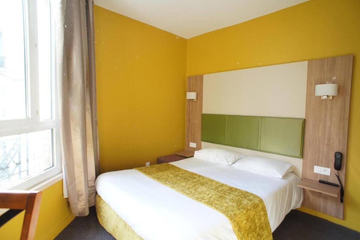 Habitació d'hotel amb un llit gran, una taula de nit, un taulell i una finestra amb cortines, i una làmpada de paret.