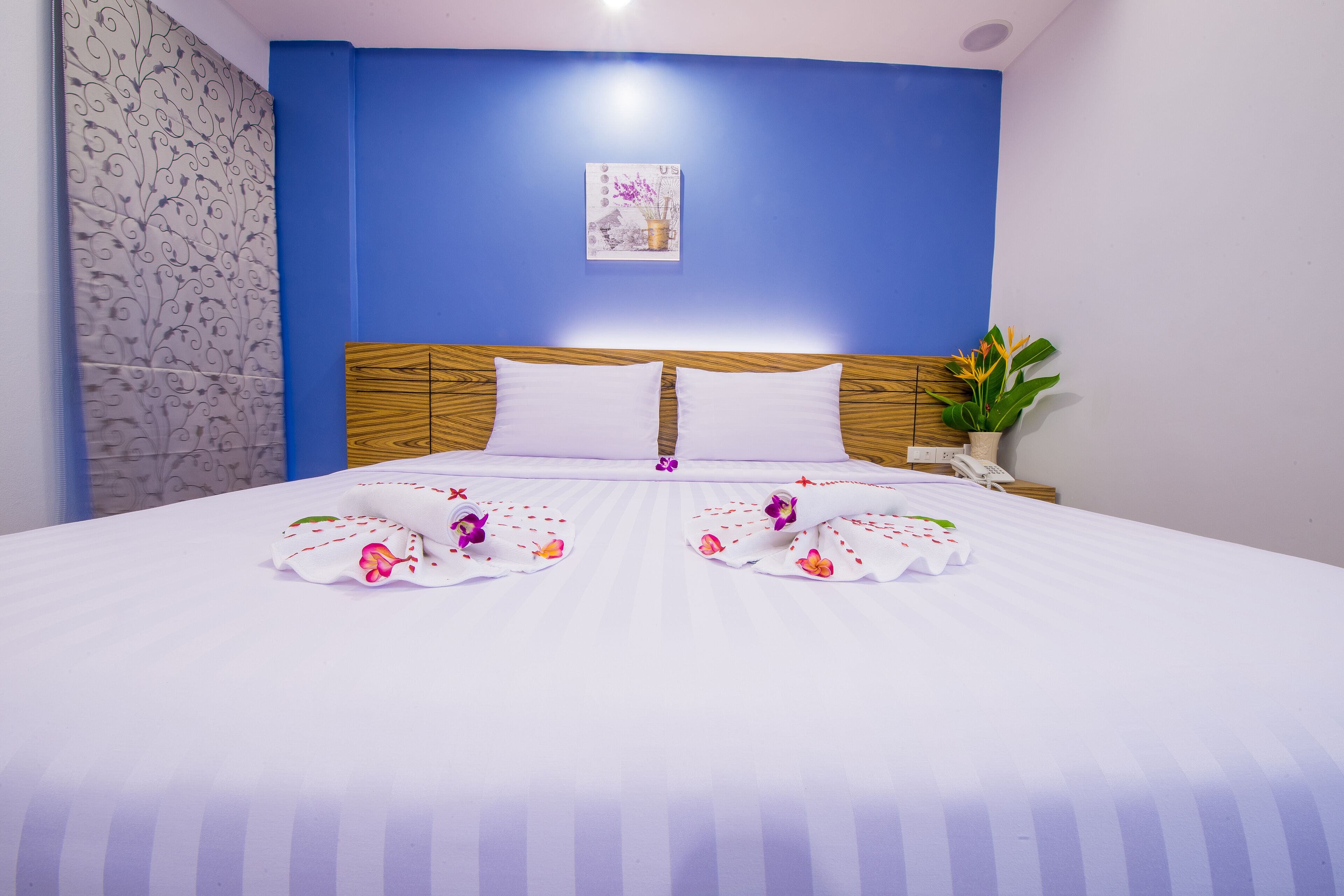 A neatly made bed with white linens and floral decorations, a blue wall, and a small plant on the side.