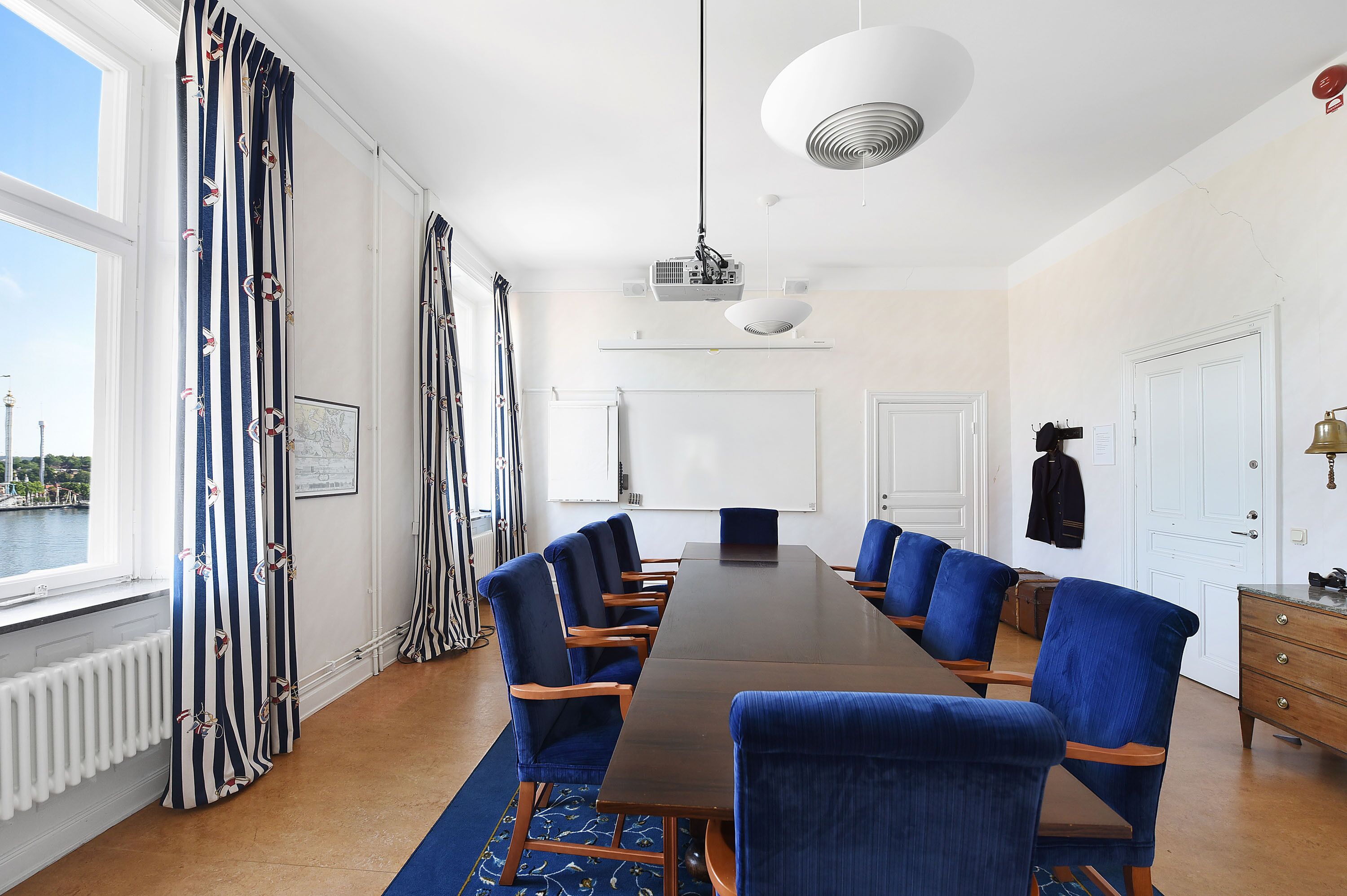 Ein Konferenzraum mit langem Tisch, blauen Stühlen, einem Projektor und Blick auf ein Gewässer durch große Fenster.