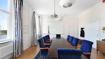 Ein Konferenzraum mit langem Tisch, blauen Stühlen, einem Projektor und Blick auf ein Gewässer durch große Fenster.