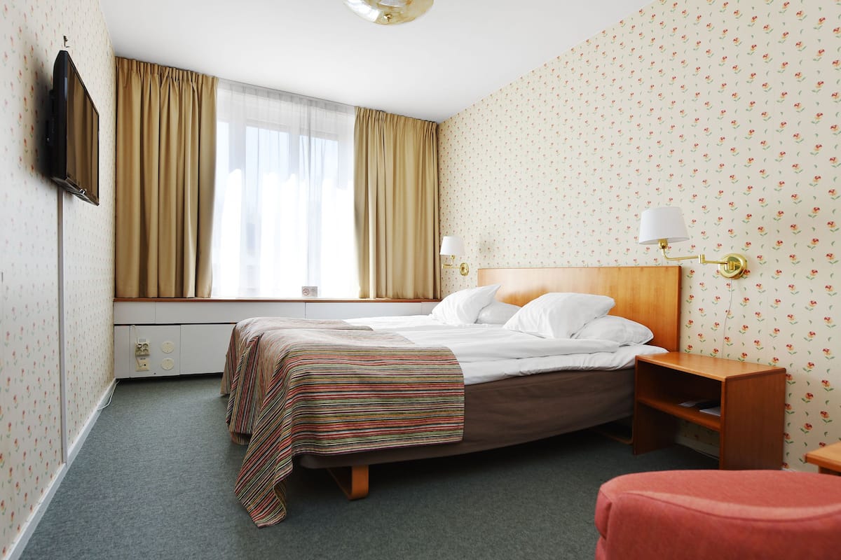 Una habitación de hotel con cama, mesita de noche, lámpara, televisor y ventana con cortinas.
