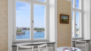 Una habitación con un ventanal que ofrece vistas a la ciudad, un radiador, una mesa con una revista y una silla.