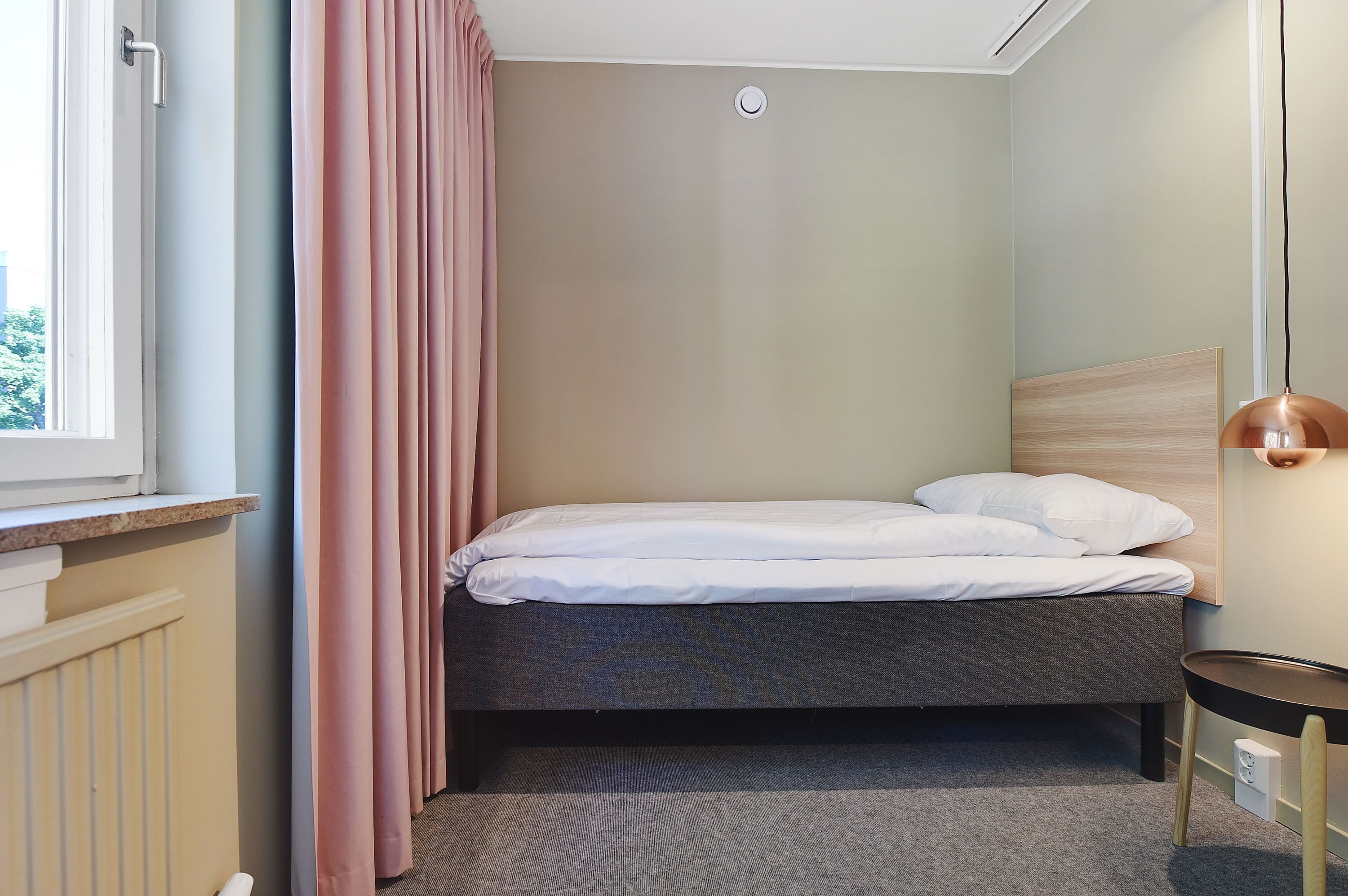 Une petite chambre à coucher, bien rangée, avec un lit simple, une fenêtre avec des rideaux, un radiateur et une table de chevet avec une lampe.