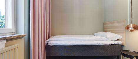 Une petite chambre à coucher, bien rangée, avec un lit simple, une fenêtre avec des rideaux, un radiateur et une table de chevet avec une lampe.