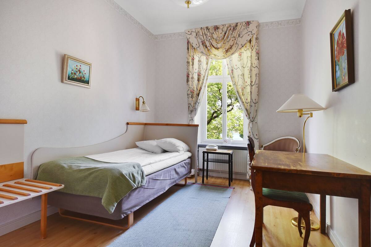 Un dormitorio con una cama individual, una mesita pequeña, una silla, un escritorio y una ventana con cortinas.