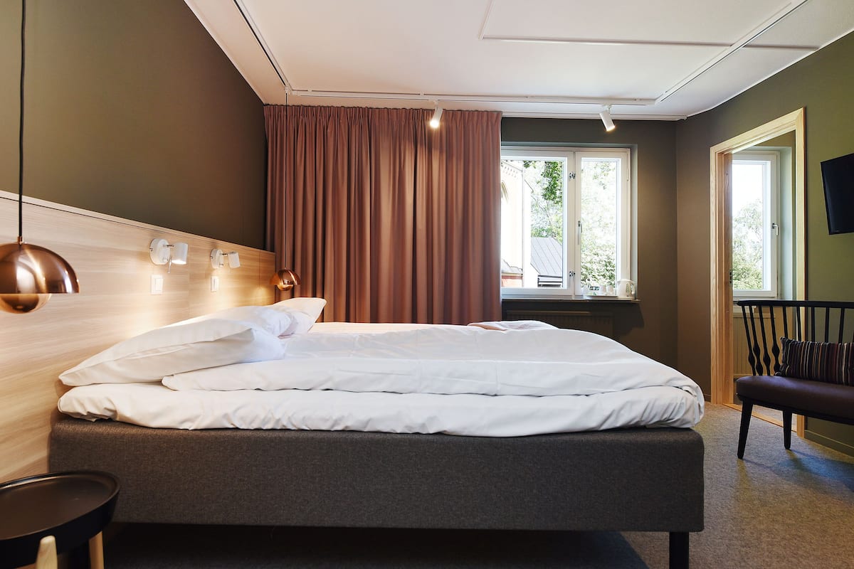 Una habitación de hotel moderna con una cama grande, un cabecero de madera, una ventana con cortinas, un televisor de pantalla plana y un banco junto a la ventana.