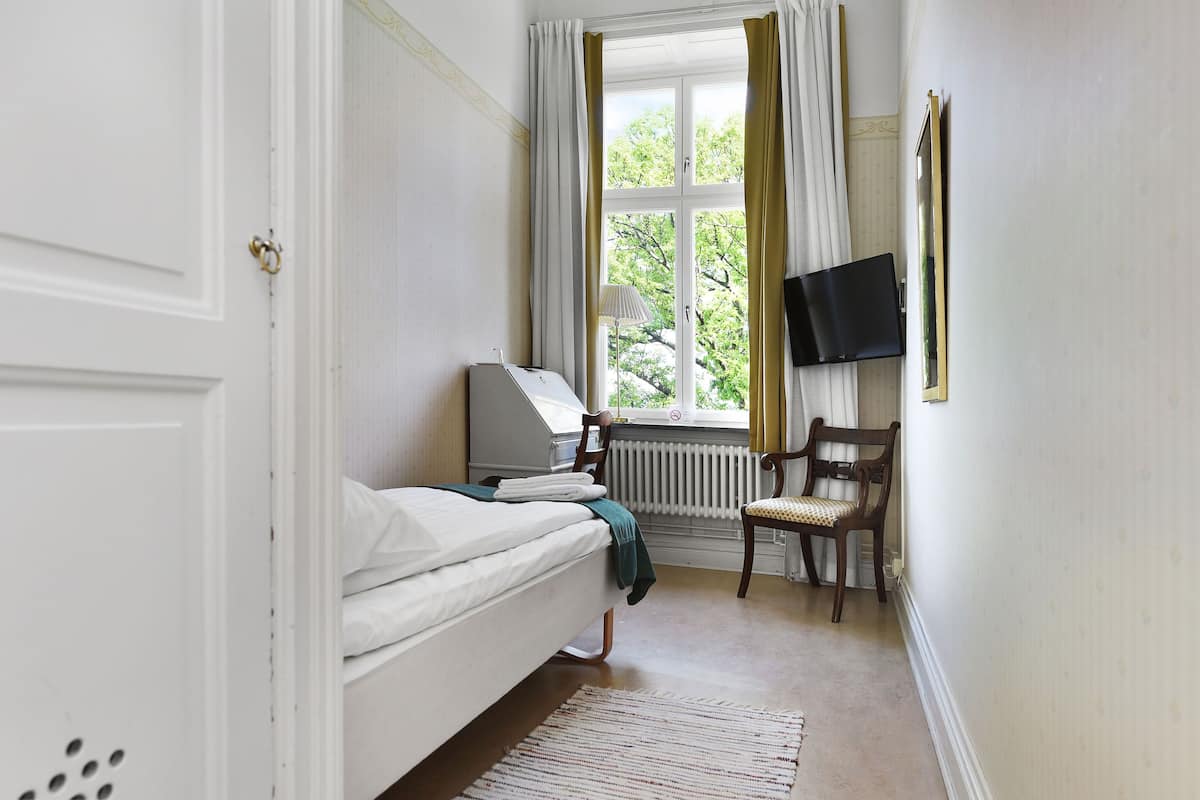 Un dormitorio con cama, escritorio con computadora, silla, televisor y ventana con cortinas.