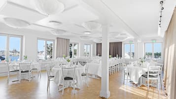 Een ruime eetruimte met meerdere ronde tafels, witte stoelen en grote ramen met uitzicht op een stadslandschap en water.