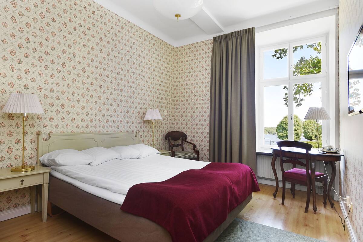 Un dormitorio con cama, escritorio y silla. Hay una ventana con cortinas, un radiador y una lámpara sobre el escritorio.