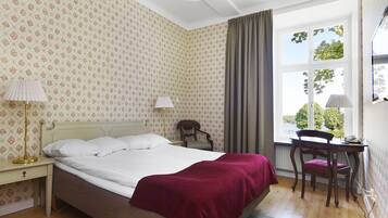 Een slaapkamer met een bed, een bureau en een stoel. Er is een raam met gordijnen, een radiator en een lamp op het bureau.