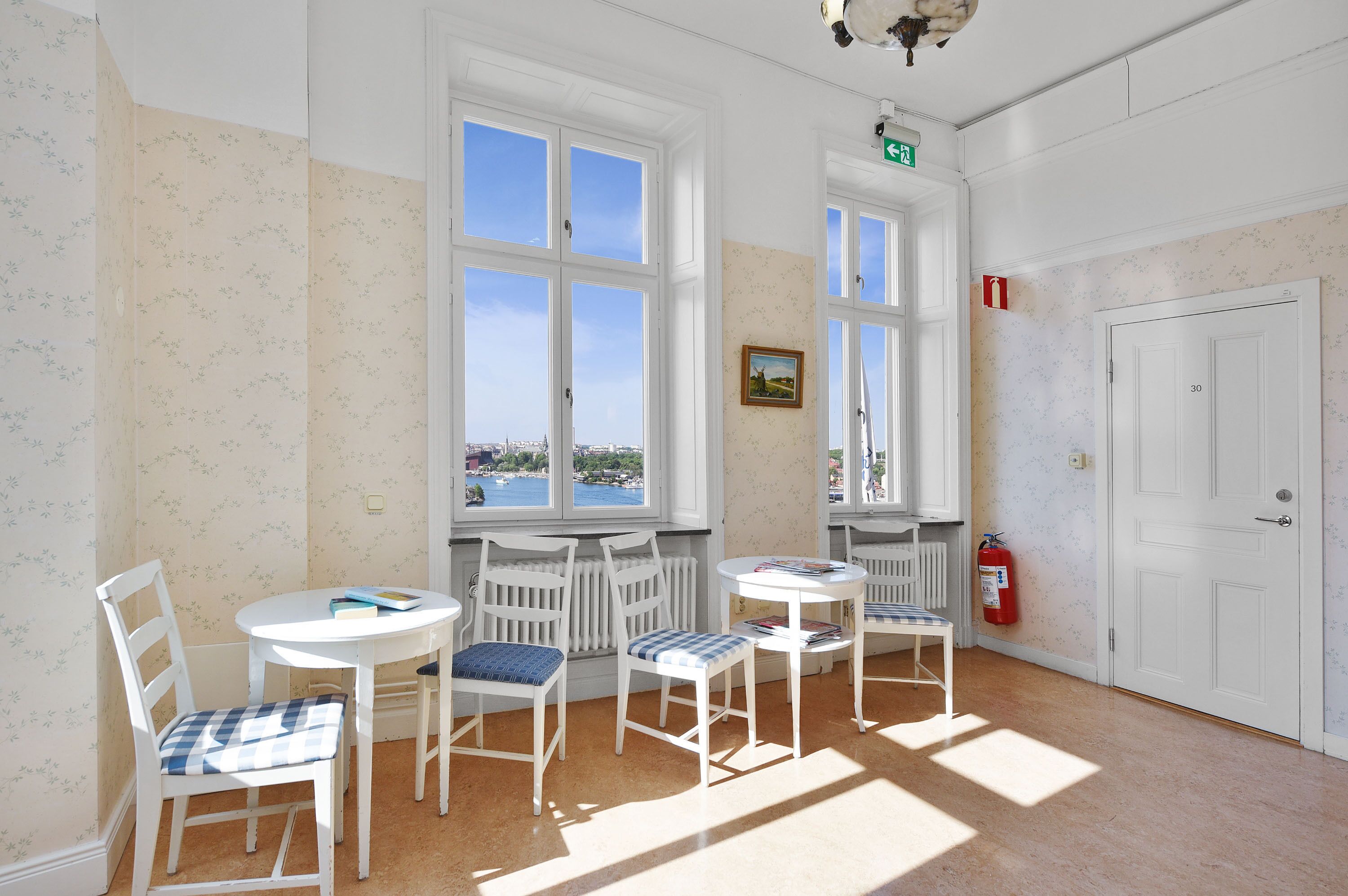 Ein Zimmer mit weißen Wänden, floralem Wandpapier und großen Fenstern. Vier weiße Stühle mit blau-weißen karierten Kissen, ein kleiner runder Tisch und ein an der Wand befestigter Feuerlöscher sind vorhanden.