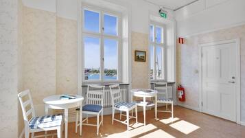 Ein Zimmer mit weißen Wänden, floralem Wandpapier und großen Fenstern. Vier weiße Stühle mit blau-weißen karierten Kissen, ein kleiner runder Tisch und ein an der Wand befestigter Feuerlöscher sind vorhanden.