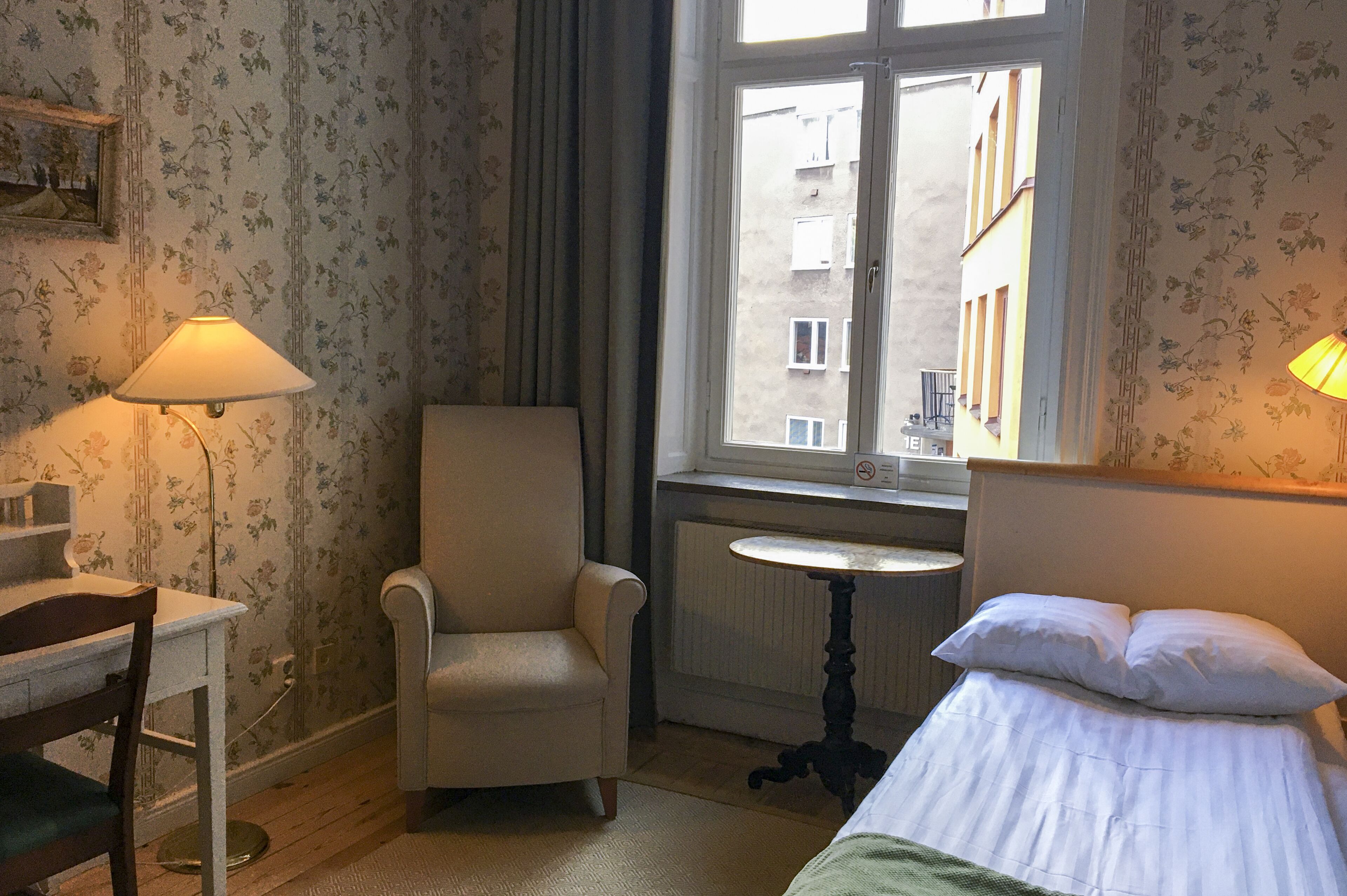 A hotel room with a bed, a chair, a small table, and a window with a view of buildings.