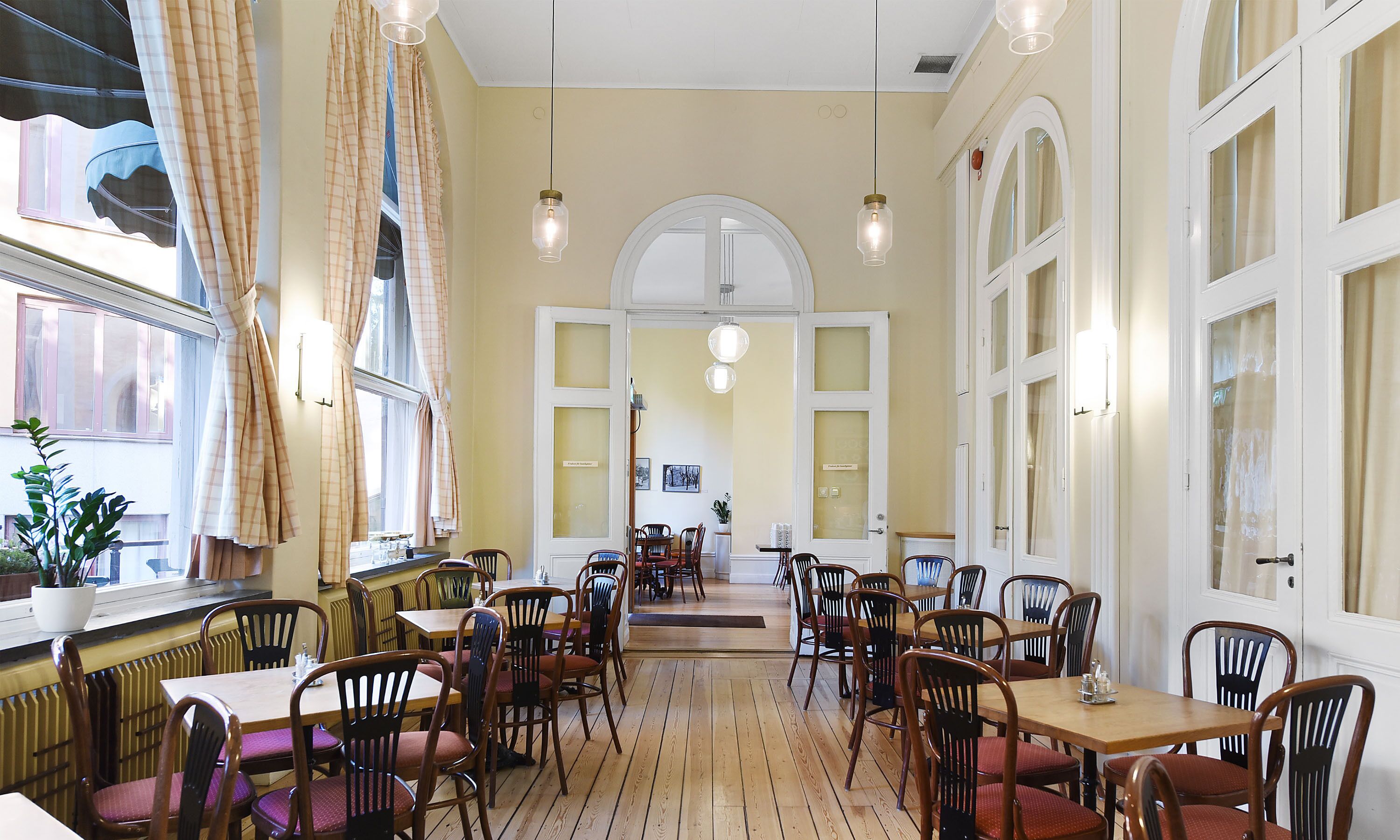A restaurant with wooden flooring, multiple tables and chairs, and large windows with curtains.