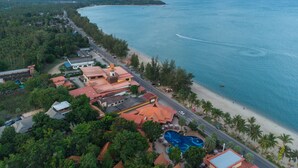 An aerial view of a coastal area with a beach, a swimming pool, and a road lined with buildings. - Baan Grood Arcadia Resort & Spa (Bang Saphan)