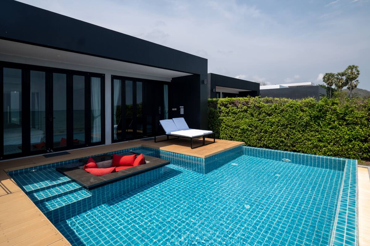 A modern outdoor pool area with a wooden deck, a lounge chair, and a small seating area with cushions.