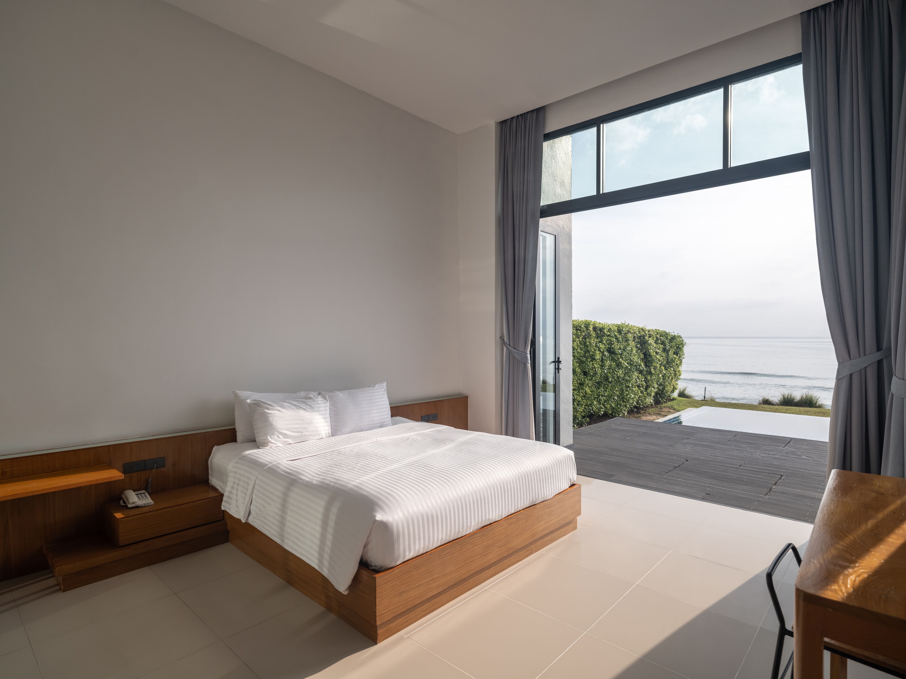A modern bedroom with a large bed, wooden bedside tables, and a view of the sea through a sliding glass door.