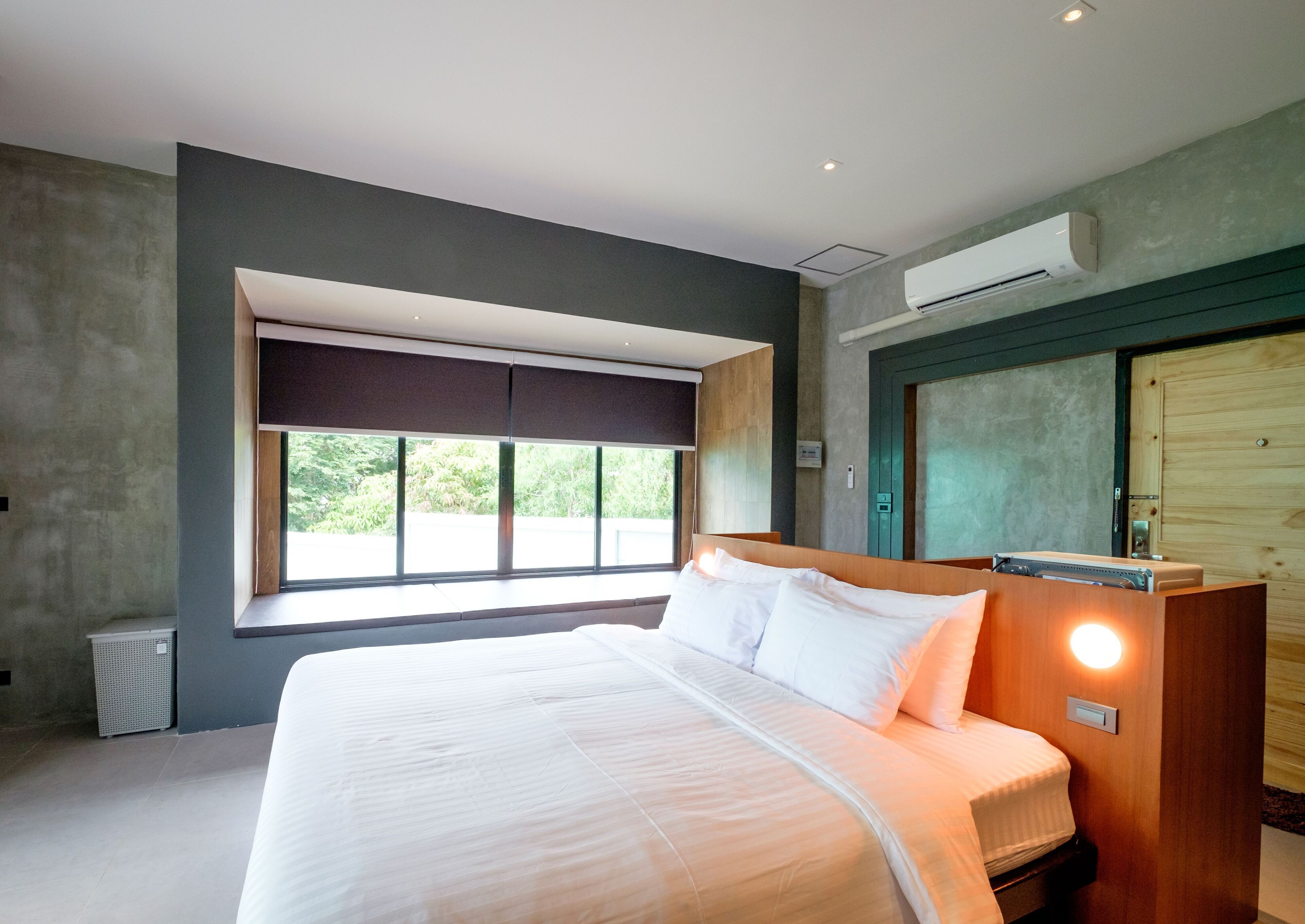 A modern hotel room with a large bed, a window with a view, an air conditioning unit, and a wooden door.