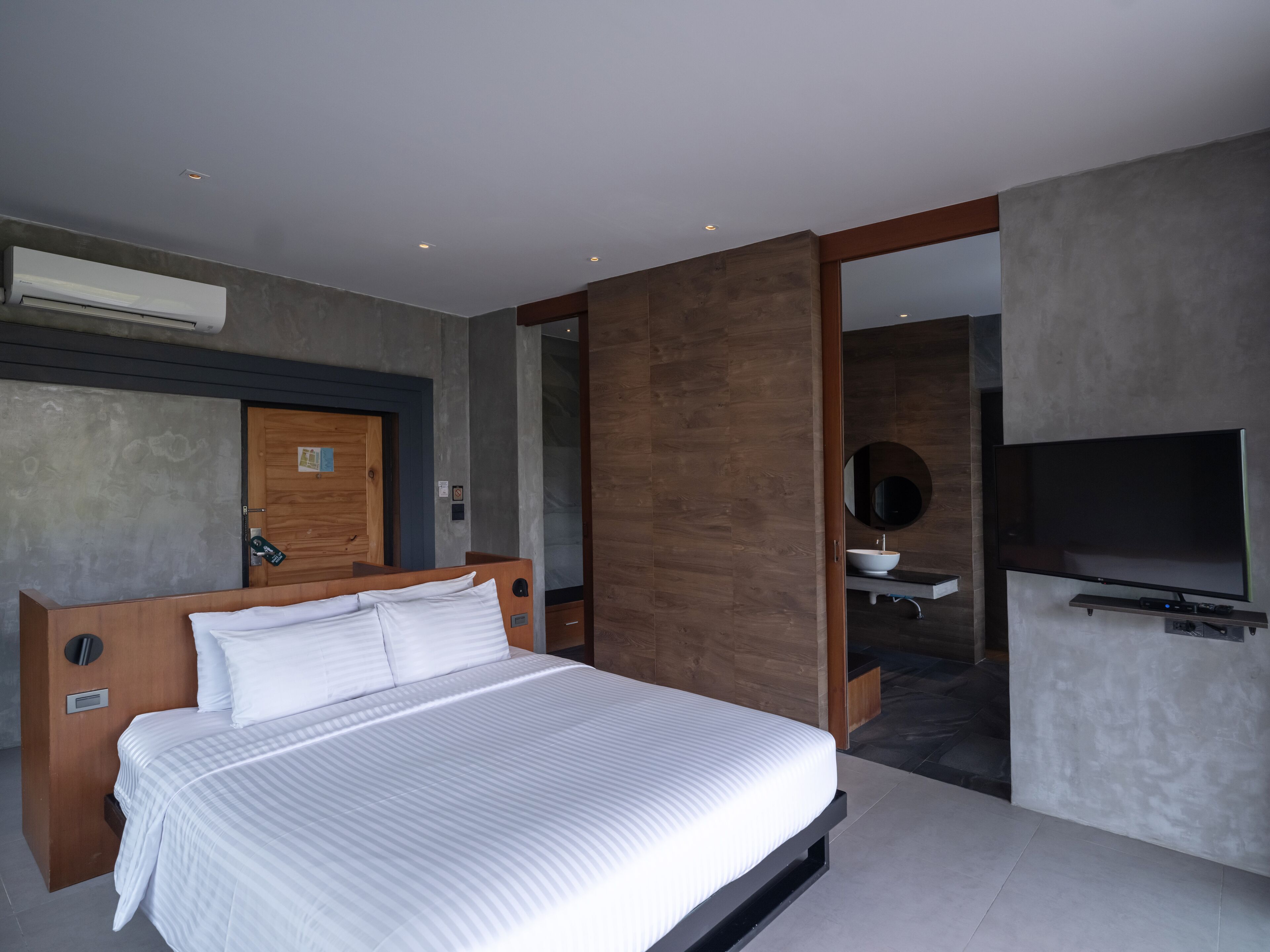 Una habitación de hotel moderna con una cama grande, una puerta de madera, un baño con lavamanos y espejo, y un televisor de pantalla plana montado en la pared.