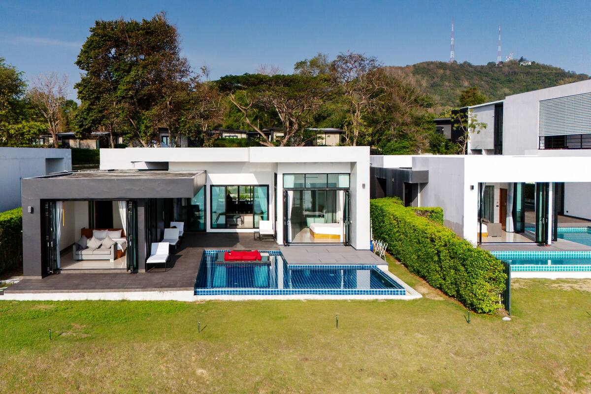 A modern house with a pool, a covered patio, and a view of a hill with trees.