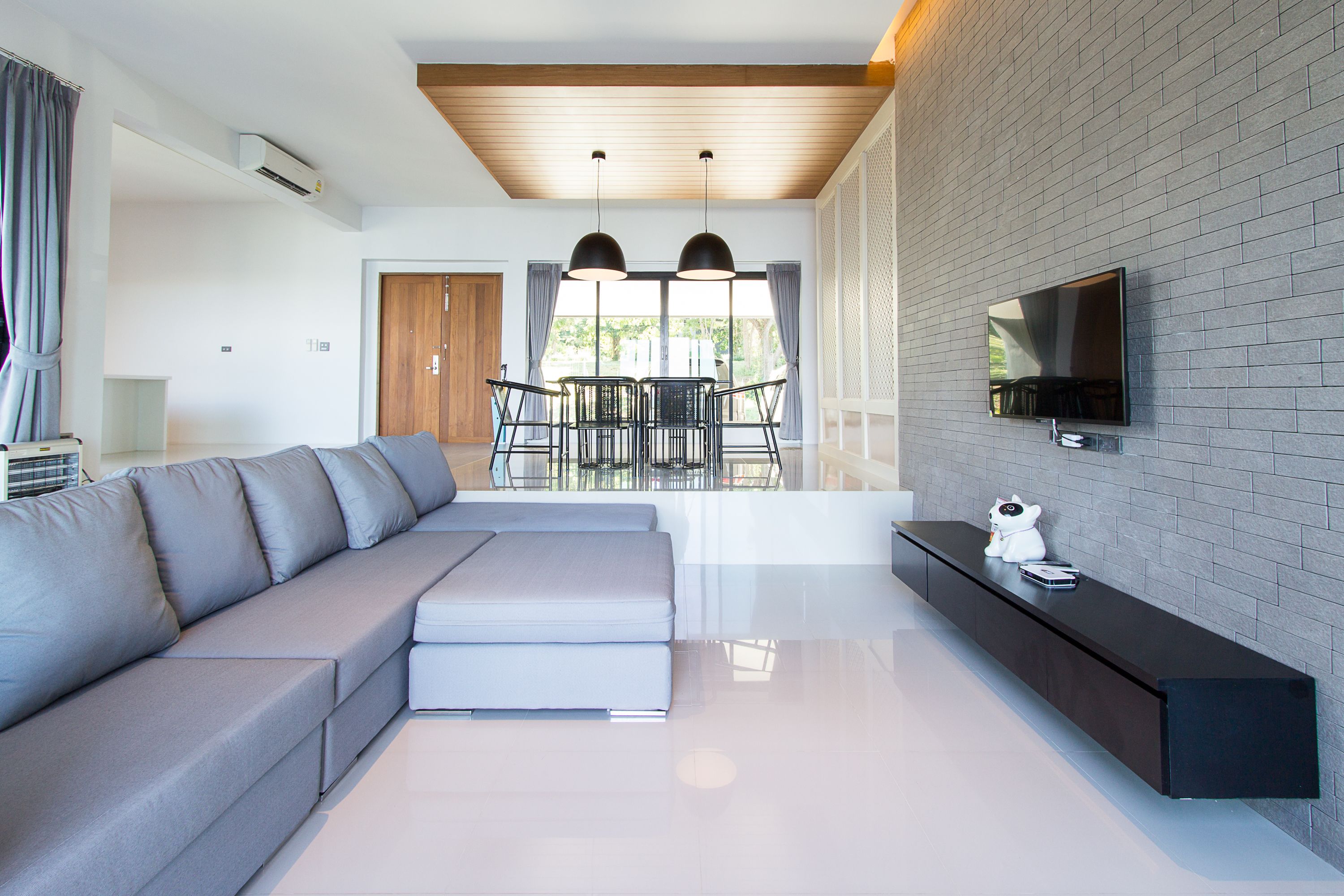 A modern living room with a grey sectional sofa, a dining area with a table and chairs, a flat-screen TV mounted on a brick wall, and a small white cat figurine on a console table.