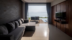 A modern living room with a large sofa, a flat-screen TV mounted on a wooden cabinet, and a view of the ocean through large windows. - Sunshine Paradise Resort (Thap Sakae)